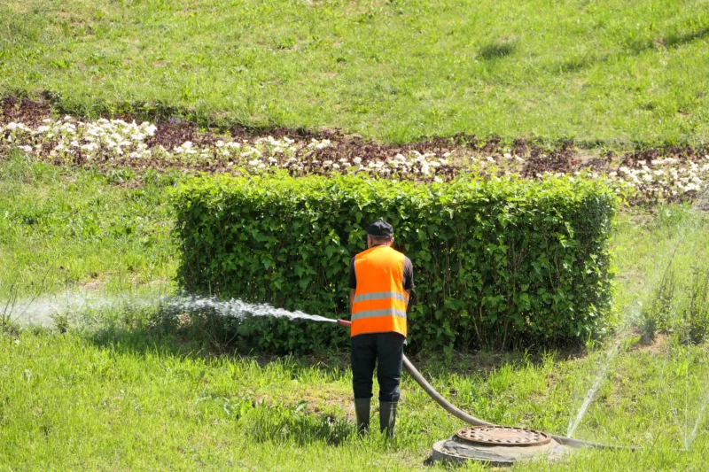 Ein Mann in Warnweste bewässert eine Grünfläche mit Wasserschlauch